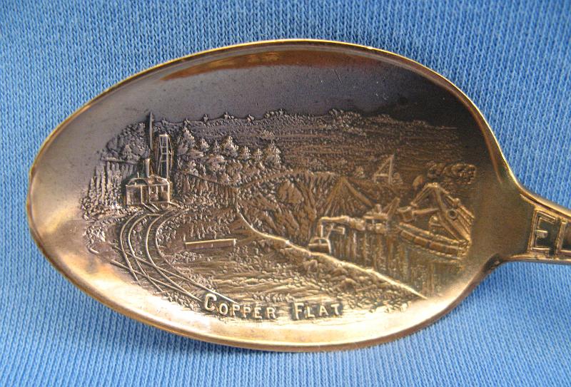 Souvenir Mining Spoon Copper Flat Bowl.JPG - SOUVENIR MINING SPOON COPPER FLAT ELY NEVADA - Copper souvenir mining spoon, embossed scene of open pit copper mine in bowl with marking COPPER FLAT at bottom, handle is a skyline depiction of mountains with buildings and smokestacks and a bridge with ELY NEVADA marked on handle, Steptoe Mining and Smelting Co. is marked at the bottom of the skyline, reverse has Paye & Baker makers mark with Sterling x’ed out, 5 1/4 in. long, weight 21 grams [Copper Flat is today a general reference to the area in eastern Nevada where the great copper discoveries were made at the first of the 20th century.  Copper Flat was also small settlement for a short time in this area (now long gone) approximately 6 miles west of Ely, NV in White Pine County.  The Copper Flat area is also associated with several small towns including Kimberly, Riepetown, Ruth and Veteran. The famed open-pit copper mines of eastern Nevada, including the Liberty Pit (largest in the state), are located in this area between Ely and Ruth just south of Highway 50. Through the first half of the 20th century, this area produced nearly a billion dollars in copper, gold and silver. The first mining claims were filed in White Pine County, Nevada, as early as 1867, and the first copper claim was filed in the summer of 1900.  Over the next couple years, an additional 26 claims covering 437 acres of the Copper Flat area were filed.  The Nevada Consolidated Copper Company was organized and incorporated under the laws of Maine on November 17, 1904 to buy up and operate these claims. By May 1906, the company was controlled by the Guggenheim family and their associates and in 1907, steam shovels began stripping the overburden above the Eureka mine to start the open pit mining operations.  In 1906, the Nevada Consolidated Copper Company and the Cumberland and Ely Mining Corporation formed a partnership to build a smelter in the area to process the ores from Copper Flat.  Named the Steptoe Valley Mining and Smelting Company, the smelter started construction in December 1906 at the small town of McGill some 12 miles north of Ely.  The smelter was completed and operations began on May 15, 1908.  The first copper was shipped from the smelter on August 7 of the same year. Kennecott Utah Copper acquired Nevada Consolidated Copper Company, which included Steptoe Valley Mining and Smelting Company and the smelter at McGill, in 1932. In 1983, the price of copper along with the low grade ore being mined led to Kennecott closing the smelter and demolishing it.]